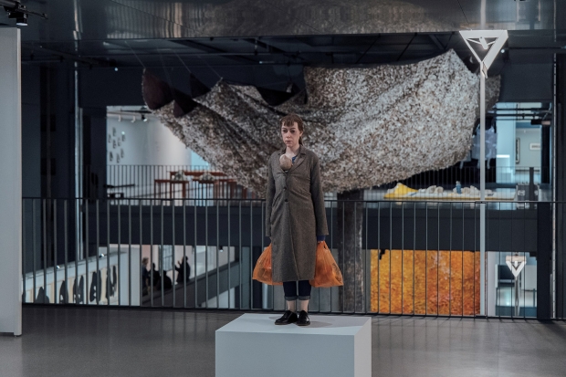 前方為澳洲藝術家Ron Mueck的作品《Woman with Shopping》；後方是《Être nature》展區內懸掛着Solange Pessoa 創作的巨型裝置藝術作品《Miracéus》，此作品由成千根鳥羽構築而成，蘊含靈性與薩滿文化精神，邀請觀眾潛入人性中原始的動物本質。（照片 © Cyril Marcilhacy）